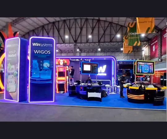 Stand de feria con iluminación de neón azul y morado, pantallas digitales, máquinas recreativas y logotipos de "Win Systems", situado dentro de una gran sala de exposiciones cubierta.