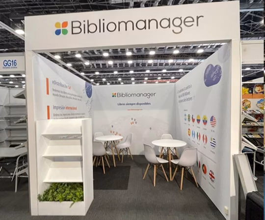 Stand de Bibliomanager en un evento con mesas, sillas, folletos y un despliegue de banderas internacionales en la pared.