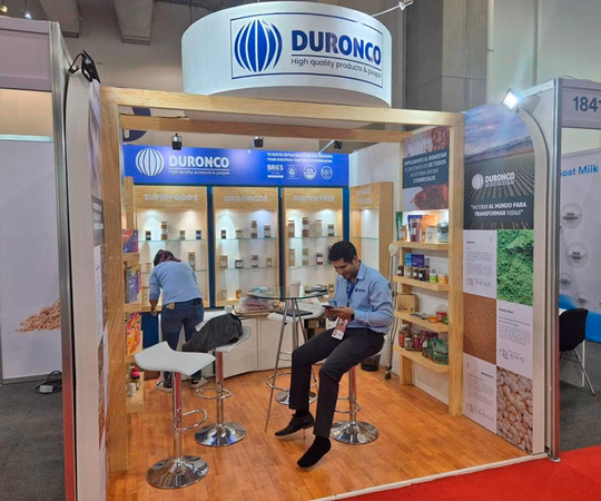 Un hombre se sienta y lee en el stand de la feria DURONCO, que muestra productos alimenticios envasados en estanterías, con otra persona ordenando los artículos al fondo.