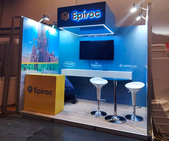 Stand de exposición con la marca Epiroc, dos taburetes de bar blancos, un mostrador amarillo, una pantalla montada en la pared y gráficos promocionales al fondo.