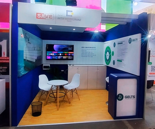 Stand de feria con logotipos de empresas, paneles informativos, una pequeña mesa redonda con dos sillas blancas y una pantalla montada en la pared sobre un suelo de madera.