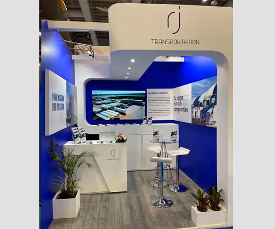 Stand de exposición con decoración en azul y blanco, un mostrador, taburetes de bar, plantas en macetas, un televisor que muestra camiones y la marca "RJ TRANSPORTATION".