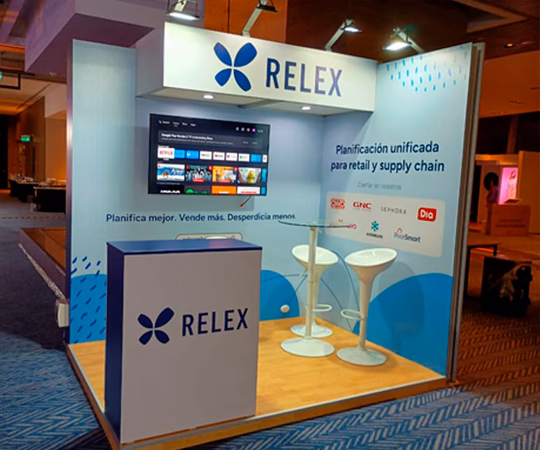 Stand de feria para RELEX con marca, dos sillas blancas, una mesa, un monitor y un telón de fondo con información sobre planificación de la cadena de suministro y venta al por menor.