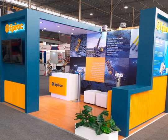 Stand de Epiroc en una feria comercial con la marca azul y amarilla, expositores informativos, una pantalla de TV, zona de asientos y material promocional dentro de una sala de exposiciones.