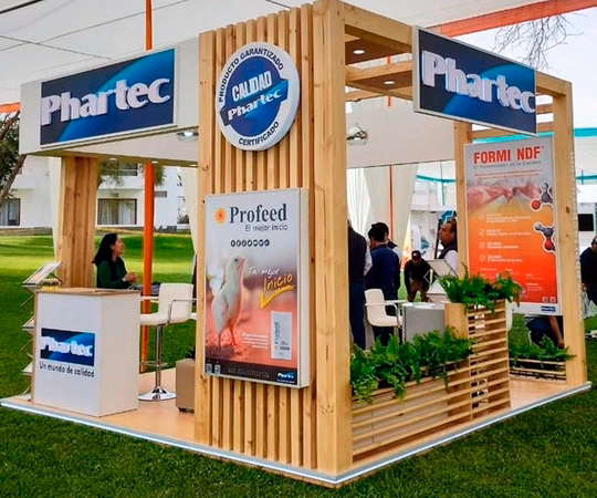 Stand de exposición de madera para PharTec con rótulos de la empresa, carteles de productos, plantas y personas interactuando dentro y alrededor del stand en un evento al aire libre.