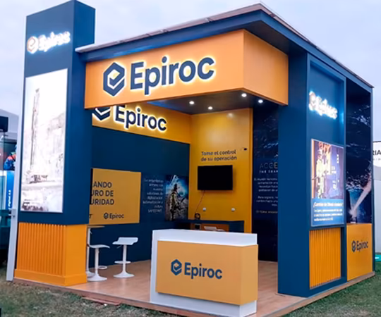 Stand de exposición de Epiroc con señalización de marca, un mostrador de recepción, dos sillas blancas, una pantalla de TV y expositores informativos.