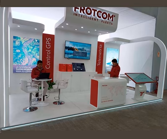 Dos personas trabajan en un stand de exposición de Frotcom en el que se muestran soluciones telemáticas y de control por GPS, con monitores, folletos y material de marca.