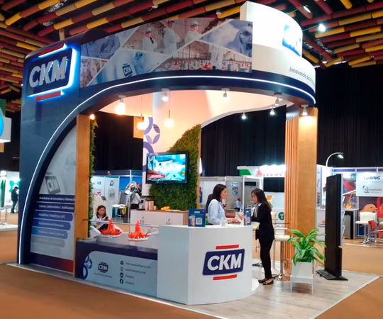Stand de exposición de CKM con la marca de la empresa, pantallas de visualización, folletos y personal interactuando con los visitantes en una feria comercial.