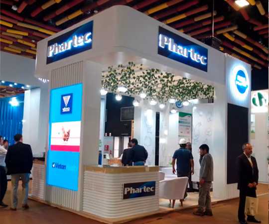 Stand de feria para Phar-tec con carteles luminosos, pantallas digitales, zona de asientos y varias personas de pie y caminando por el espacio de exposición.