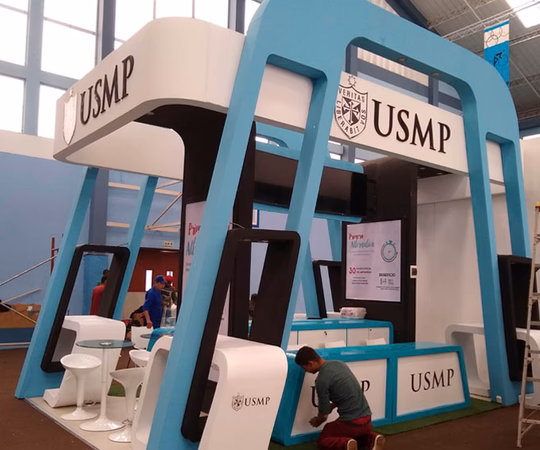 Un stand de exposición moderno con la marca USMP, detalles en azul y blanco, sillas y dos personas montando el expositor en un recinto interior.