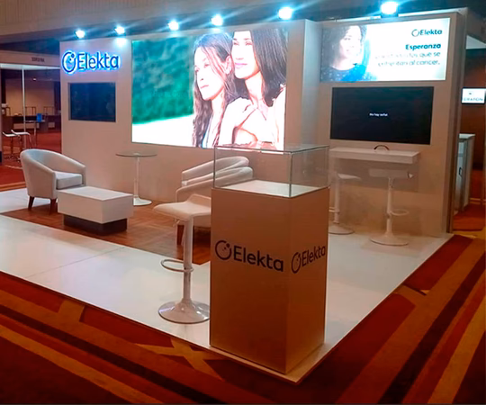 Stand de exposición de Elekta con asientos, pantallas que muestran contenido promocional, una vitrina de cristal y señalización de marca en un espacio interior para eventos.
