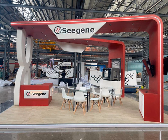 Un moderno stand de feria para Seegene con colores rojo y blanco, un mostrador de recepción, sillas, mesas y monitores de exposición dentro de una gran sala de exposiciones.