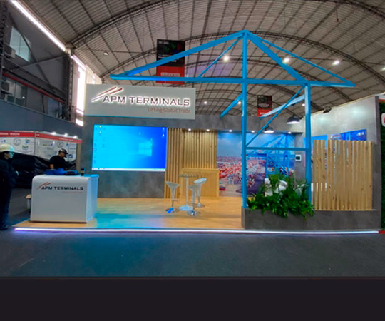 Stand de exposición de APM Terminals con mostrador de recepción, zona de asientos, gran pantalla y plantas decorativas bajo una estructura de techo curvo en el interior de un centro de convenciones.